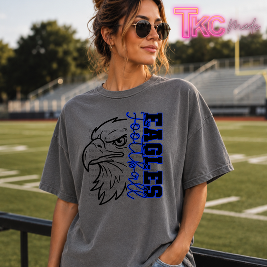 A woman wearing a gray graphic t-shirt with a black eagle head illustration and blue 'EAGLES Football' text, standing on a football field. A 'TKC Made' watermark is visible in the top right corner.