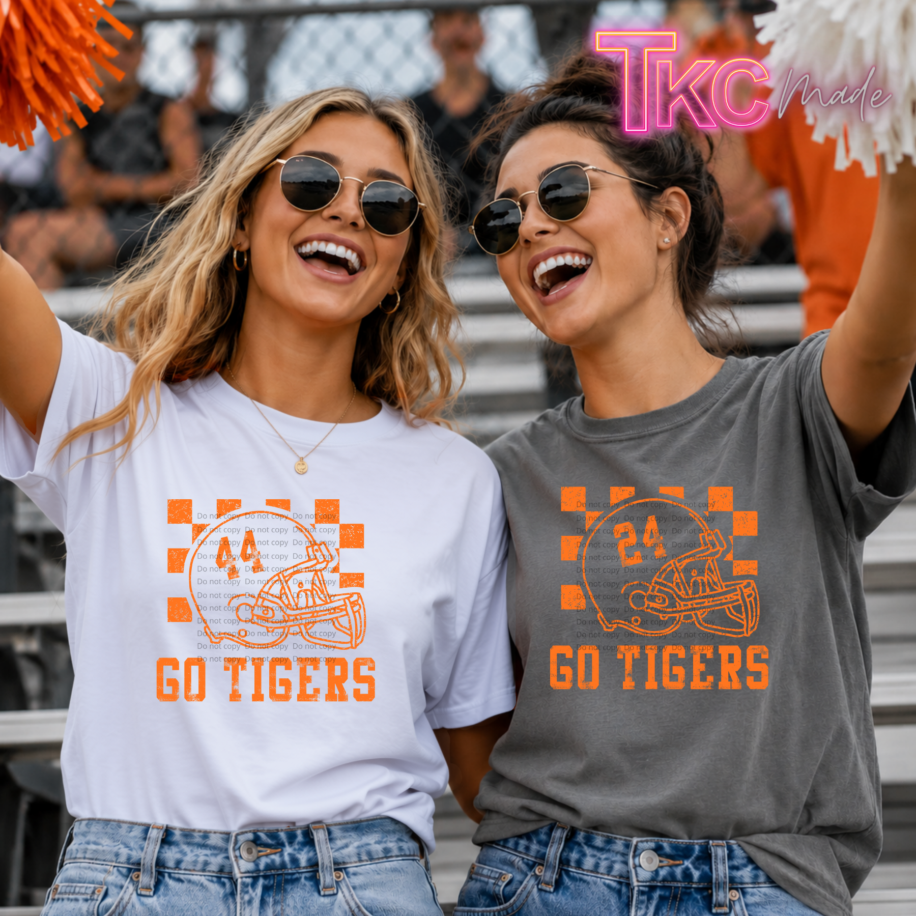 Two women wearing matching graphic T-shirts in a stadium setting. The woman on the left wears a white shirt and the woman on the right wears a dark gray shirt, both featuring an orange football helmet graphic with the number 24, checkered accents, and the text 'GO TIGERS'.