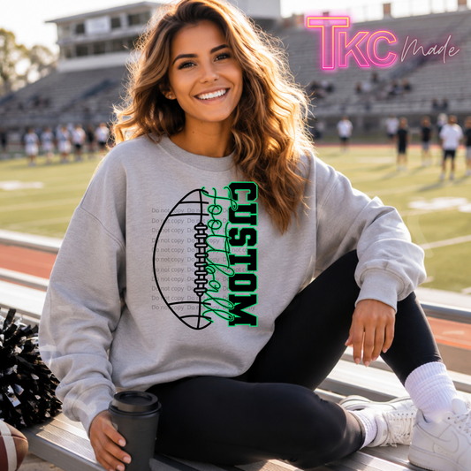A woman sitting on stadium bleachers at a football field wearing a light gray crewneck sweatshirt with a custom football graphic. The graphic features a black football outline with the word 'CUSTOM' written vertically in green and white script.