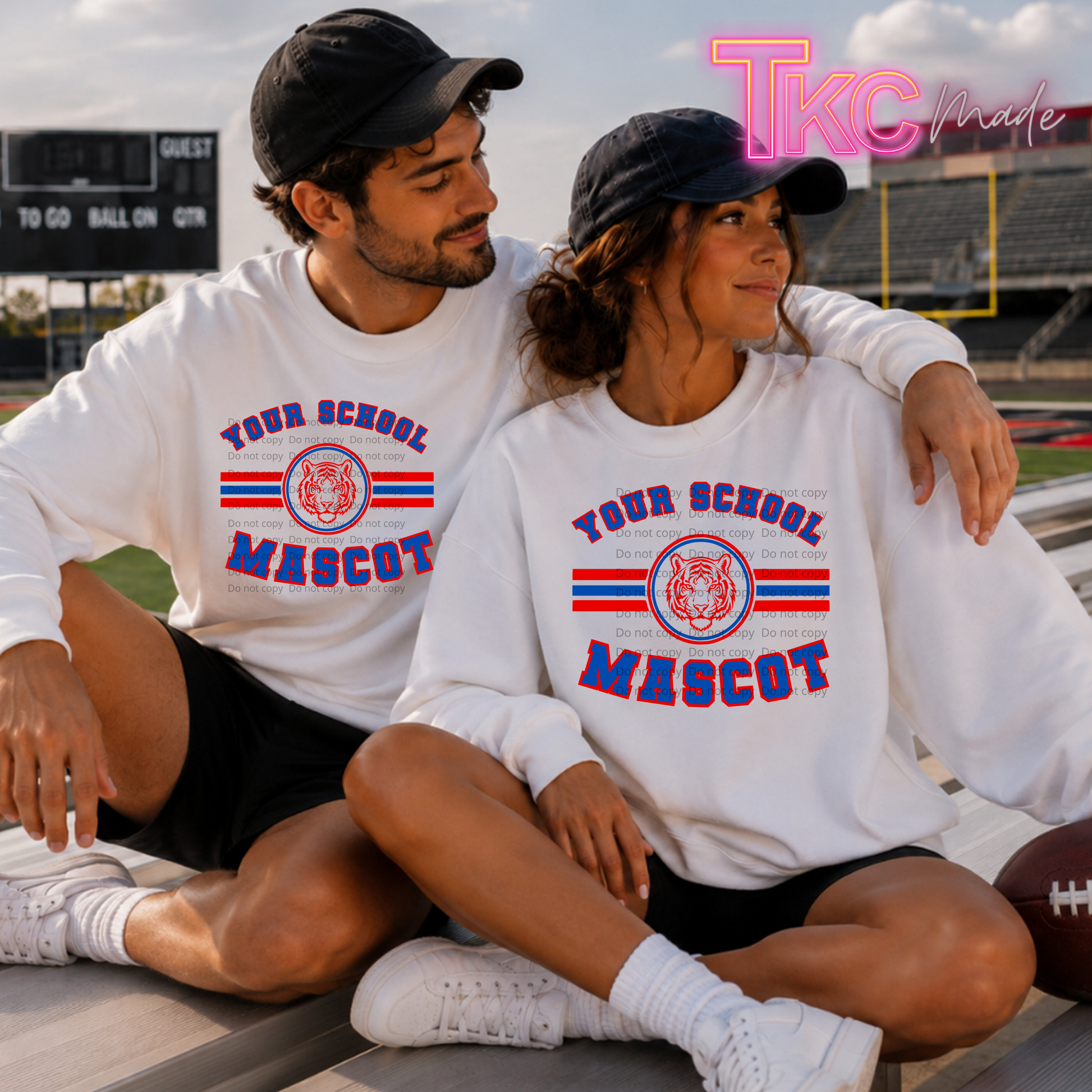 A man and a woman sitting together on outdoor bleachers wearing matching white long-sleeve sweatshirts with a red and blue 'YOUR SCHOOL MASCOT' graphic and a tiger emblem. The man wears a black cap and black shorts, and the woman wears a black cap, black shorts, and white sneakers.