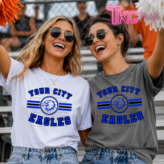 Two women in sunglasses cheering in stadium bleachers, wearing matching custom graphic t-shirts. The woman on the left wears a white t-shirt with blue and black graphics reading 'YOUR CITY EAGLES' and the woman on the right wears a heather gray t-shirt with the same blue and black graphics.