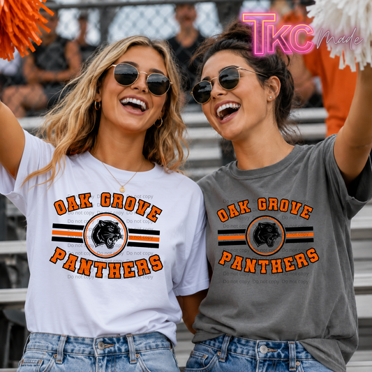 Two women wearing matching Oak Grove Panthers graphic t-shirts in white and gray, standing together in a stadium crowd. The white shirt features orange and black lettering with a panther logo, and the gray shirt is similar.