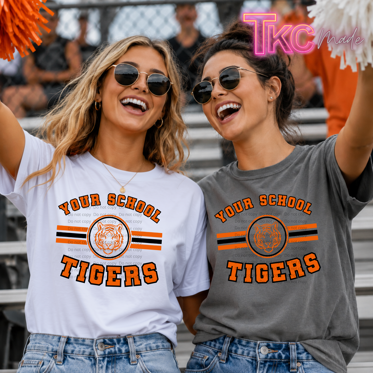 Two women wearing matching graphic t-shirts with 'YOUR SCHOOL TIGERS' and a tiger logo, standing in stadium bleachers. One shirt is white and the other is heather gray, both featuring orange and black stripes.