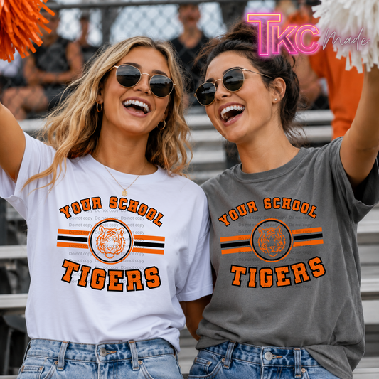 Two women wearing matching graphic t-shirts with 'YOUR SCHOOL TIGERS' and a tiger logo, standing in stadium bleachers. One shirt is white and the other is heather gray, both featuring orange and black stripes.