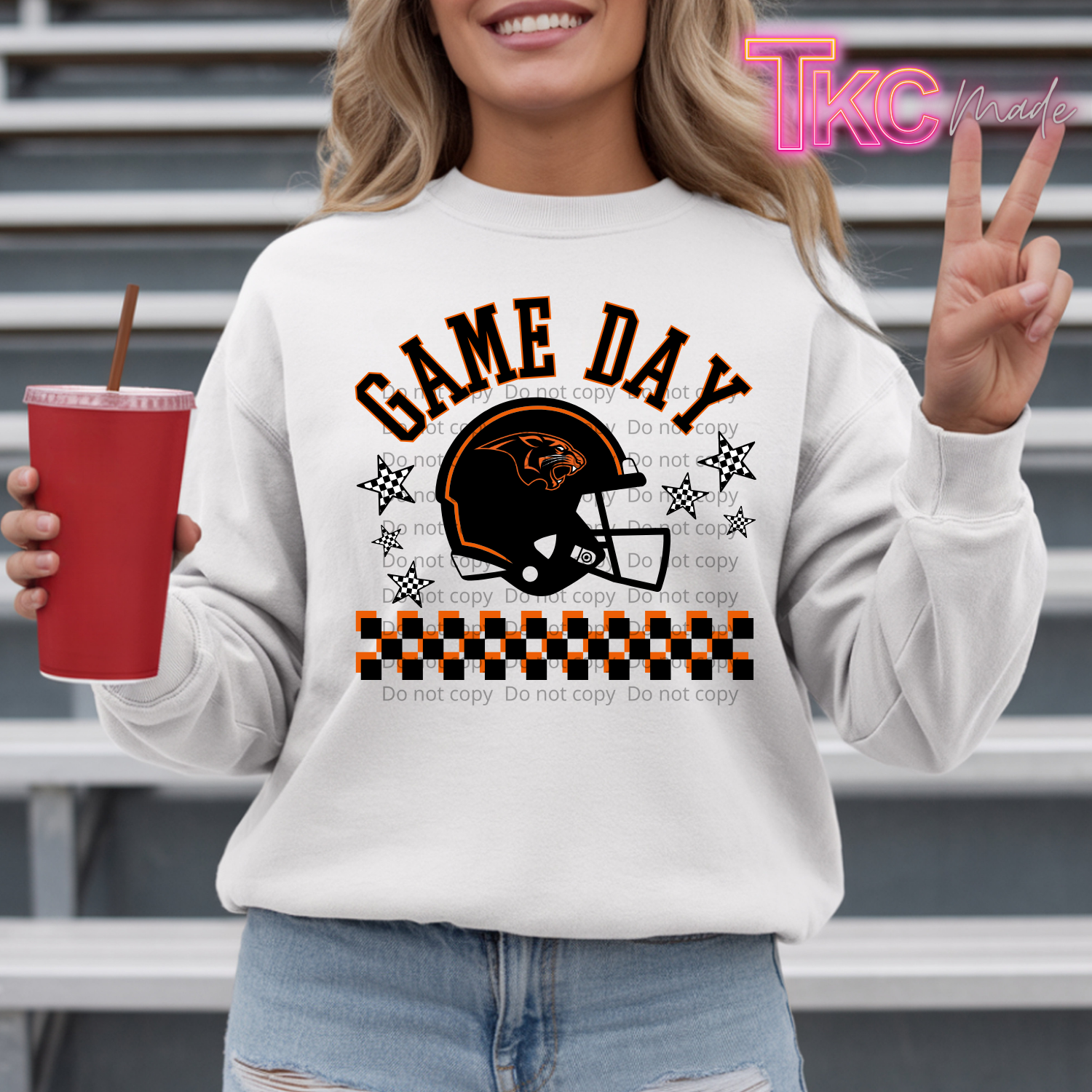 A woman wearing a white crewneck sweatshirt with a 'Game Day' football helmet graphic in orange and black. She is holding a red cup and posing in front of gray metal bleachers.