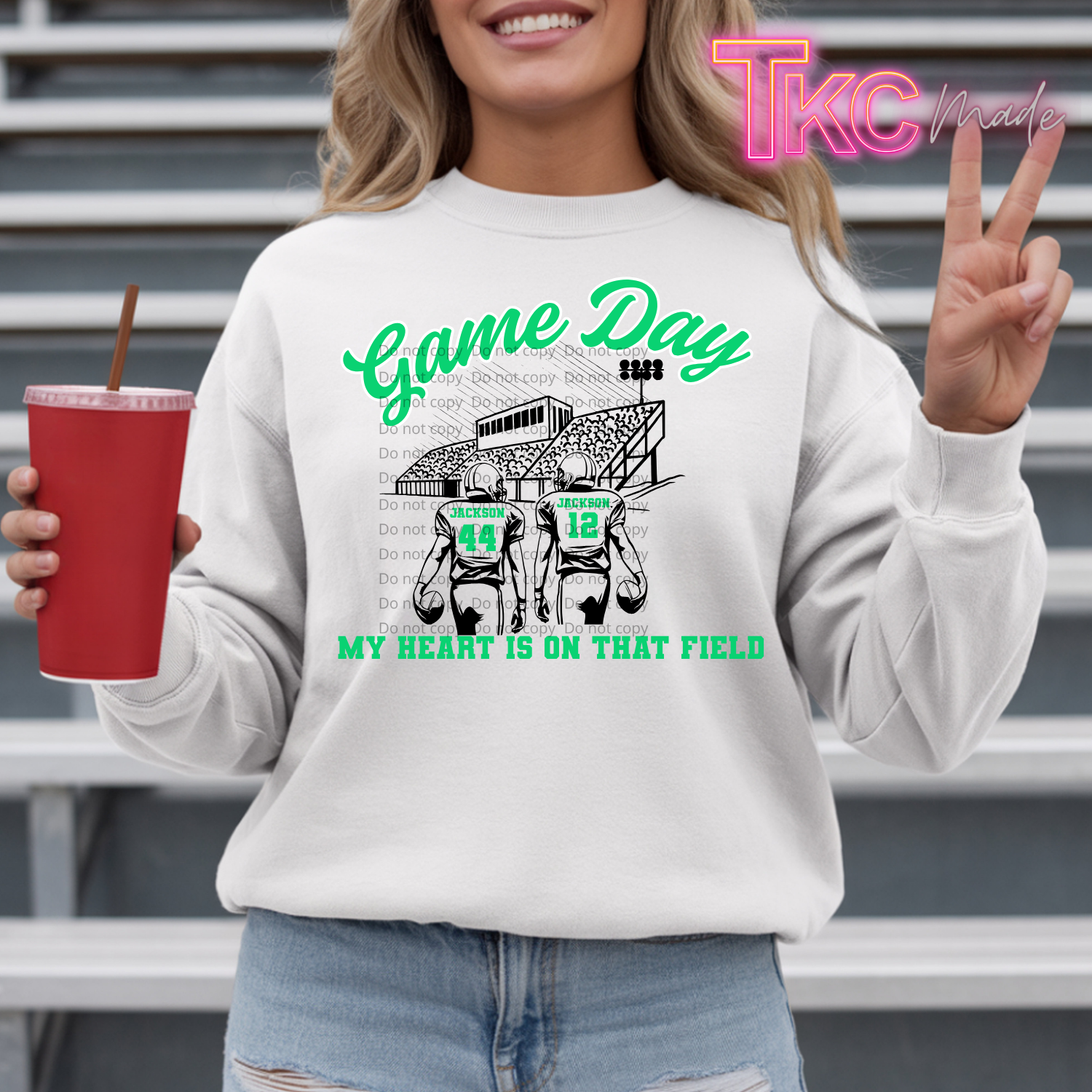 A woman wearing a white crewneck sweatshirt with a green and black football graphic that reads 'Game Day' and 'MY HEART IS ON THAT FIELD'. She is holding a red cup and making a peace sign, standing in front of metal bleachers.