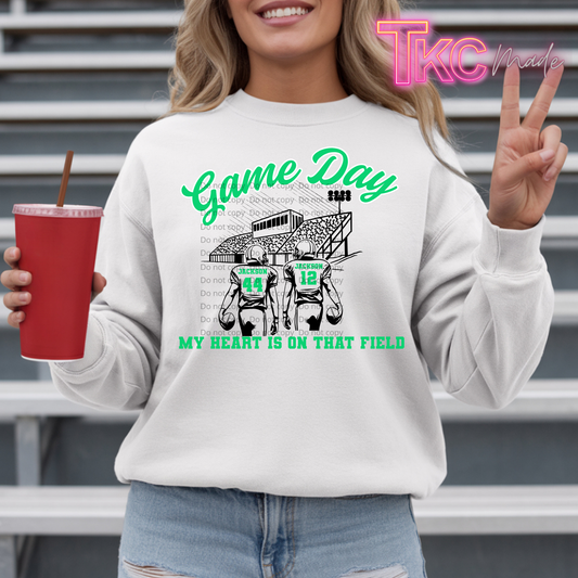 A woman wearing a white crewneck sweatshirt with a green and black football graphic that reads 'Game Day' and 'MY HEART IS ON THAT FIELD'. She is holding a red cup and making a peace sign, standing in front of metal bleachers.