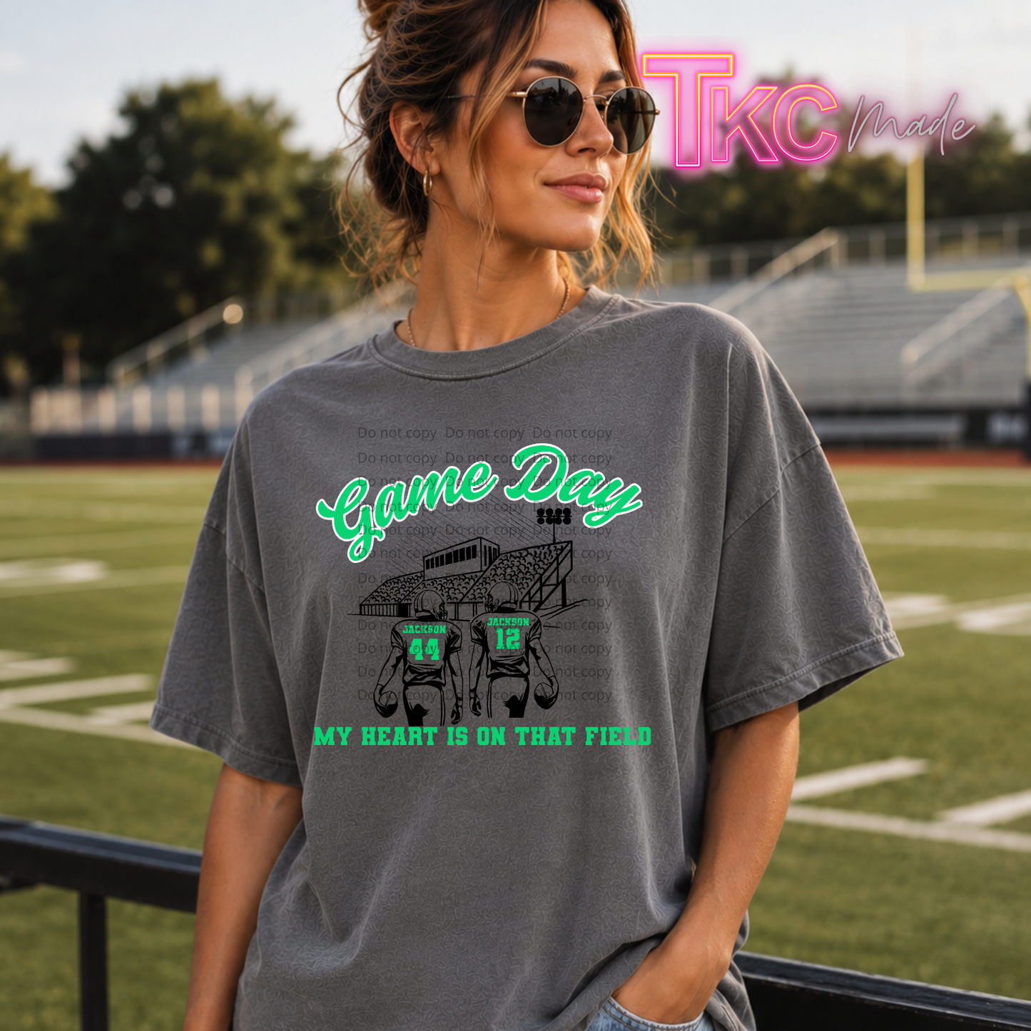A woman wearing a gray graphic t-shirt with a green and black football field design, standing on a sports field with stadium bleachers in the background. A 'TKC Made' watermark is visible in the top right corner.