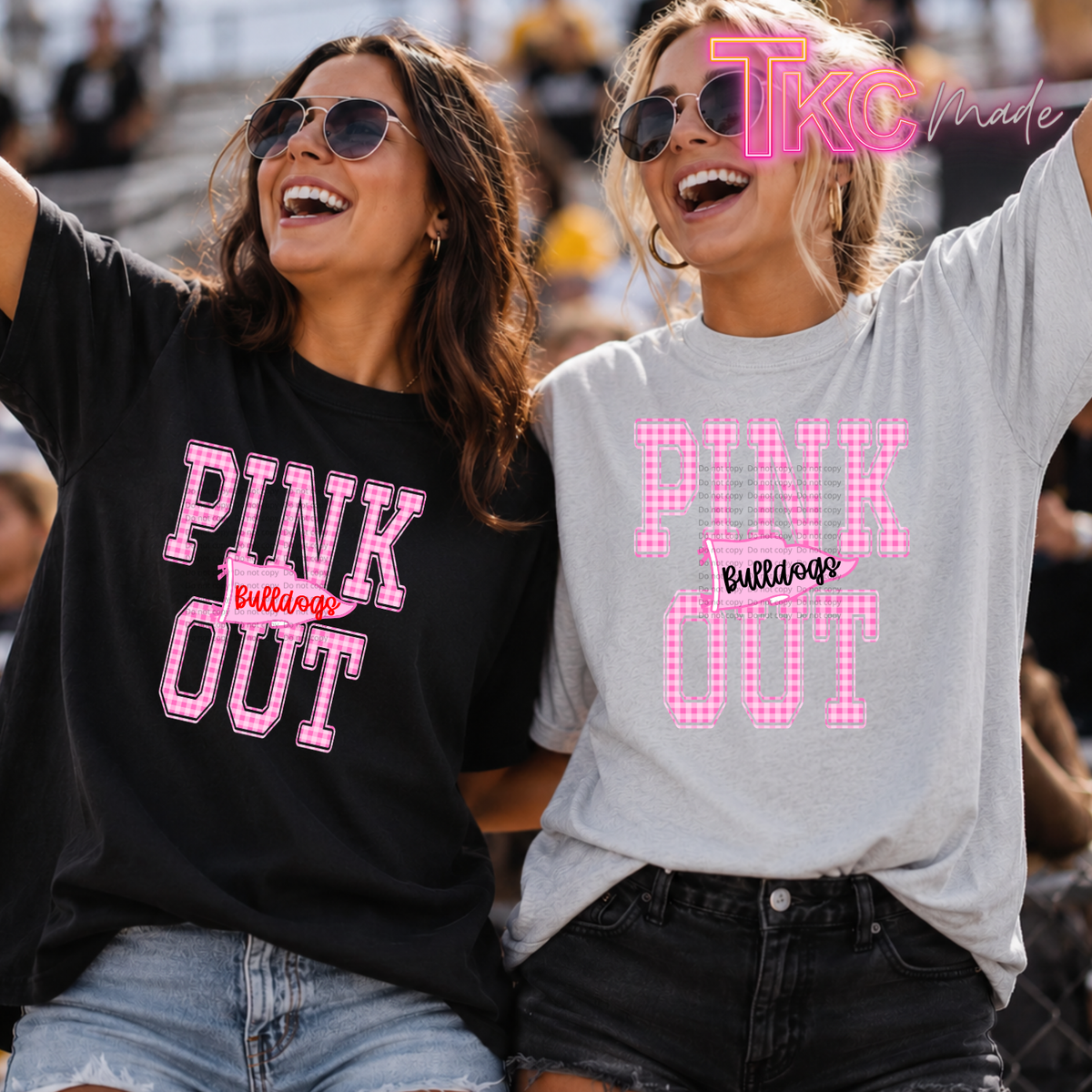 Two women at a stadium wearing matching 'PINK OUT Bulldogs' graphic t-shirts, one black and one heather gray, with pink gingham lettering.