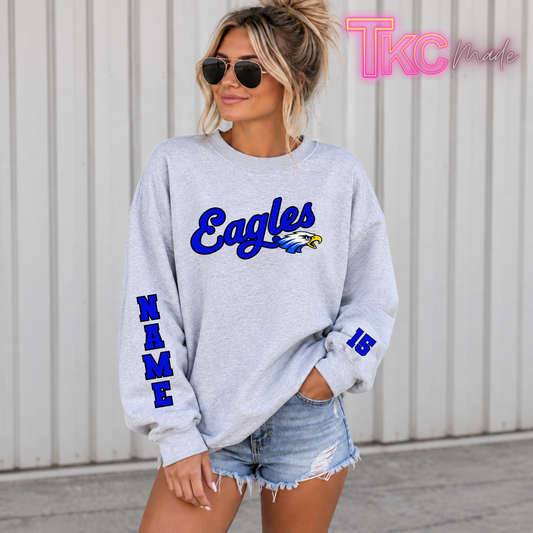 A woman modeling a custom heather gray crewneck sweatshirt featuring a blue and black "Eagles" logo with an eagle head graphic on the chest, vertical custom blue and black name lettering down the right sleeve, and a blue jersey number 15 on the left cuff, paired with distressed denim shorts.