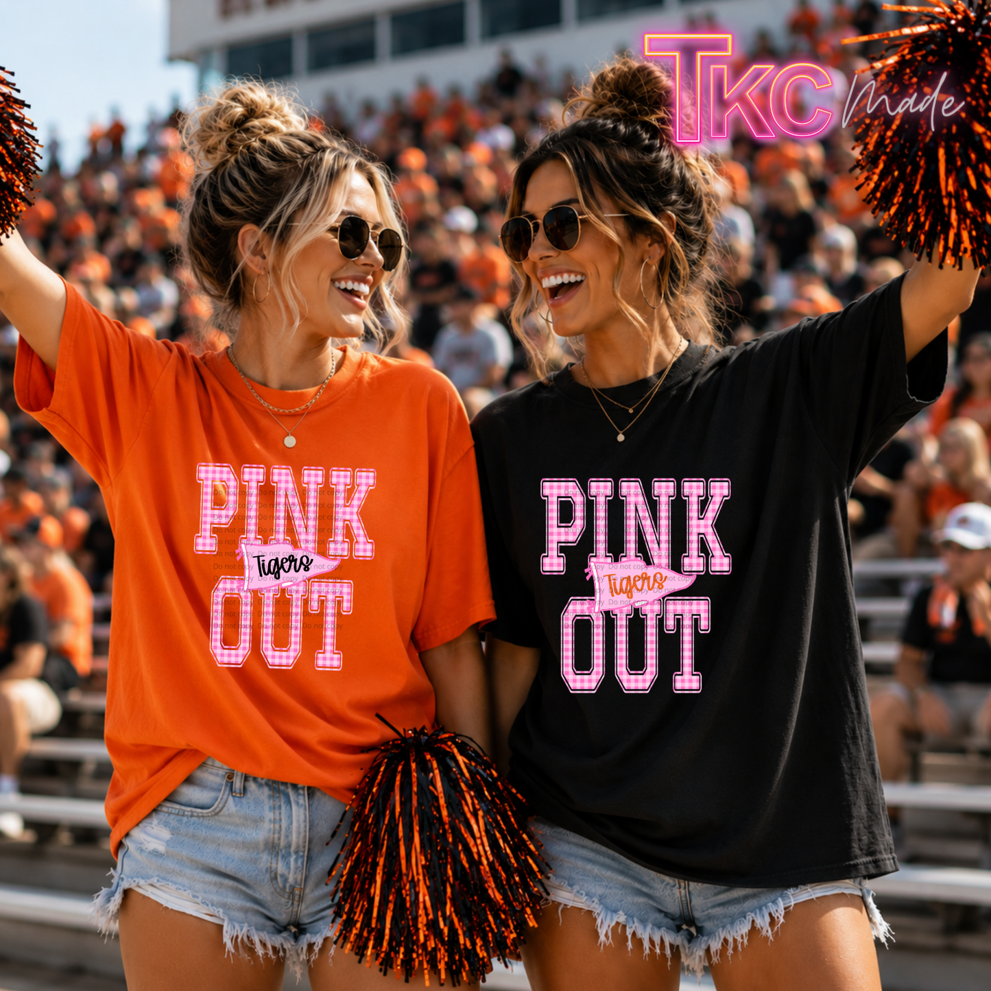 Two women in a stadium wearing oversized graphic T-shirts that read 'PINK OUT' with a pennant flag that reads Tigers. One shirt is bright orange and the other is black, both with pink outlined lettering.