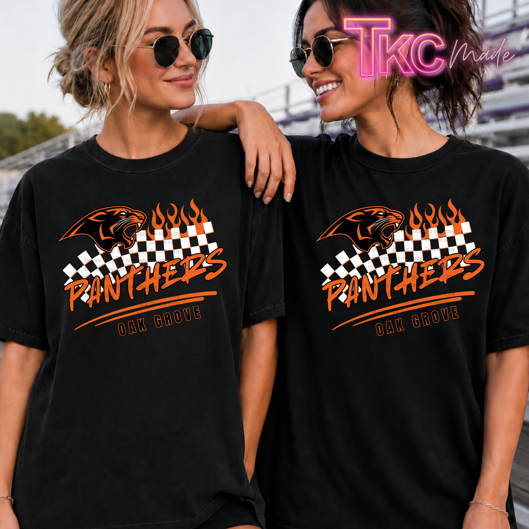Two women wearing matching black graphic T-shirts with an orange and white panther design, checkered stripe, and the text 'PANTHERS OAK GROVE'. The photo is taken outdoors with stadium seating in the background, and a 'TKC Male' watermark is visible in the top right corner.