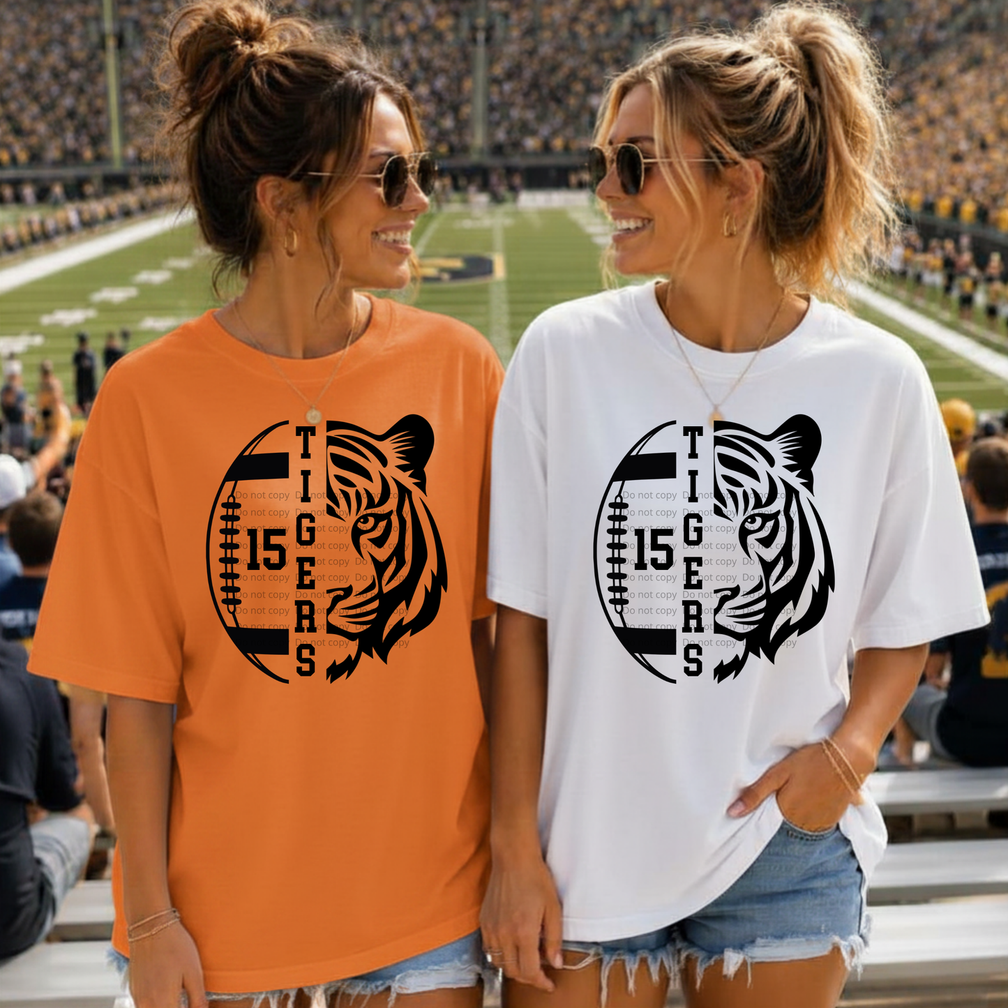 Two women standing on a football field wearing matching graphic t-shirts. The woman on the left wears an orange t-shirt with a black tiger and football graphic, and the woman on the right wears a white t-shirt with the same graphic.