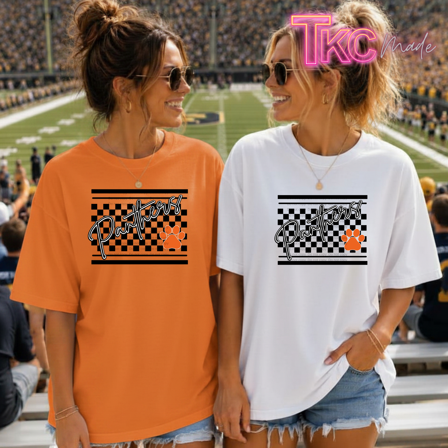 Two women standing in a stadium wearing matching graphic t-shirts. The woman on the left wears an orange t-shirt and the woman on the right wears a white t-shirt.
