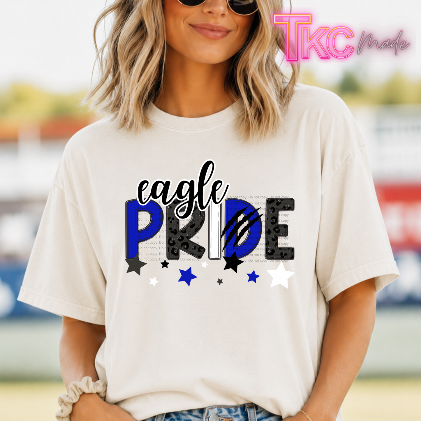 A woman wearing a cream-colored graphic t-shirt with 'eagle PRIDE' printed on the front in black, white, and blue. The background is a blurred outdoor setting.